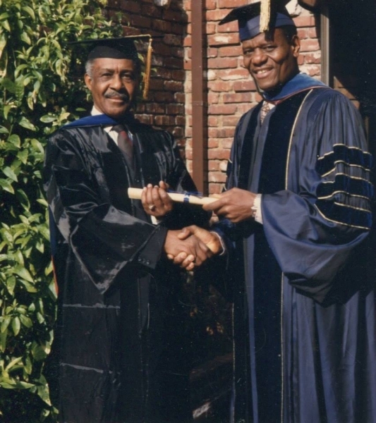 Two men in doctoral regalia shaking hands over a diploma scroll outdoors
